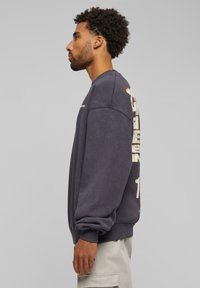 Sudadera gris con mangas largas, cuello redondo y diseño de logo lateral en dorado y blanco, con un corte relajado y textura suave.