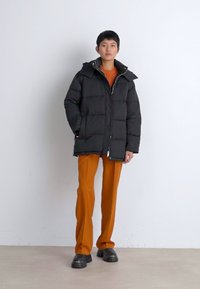 Svart pufferjakke med glidelås foran, med hette og quiltet design. Kombinert med oransje bukser og kraftige svarte sko.