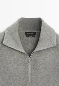 Grauer Baumwollpullover mit einem strukturierten hohen Kragen und einem silbernen Reißverschluss, der zur Hälfte vorne heruntergezogen ist und das Markenlabel innen am Kragen zeigt.