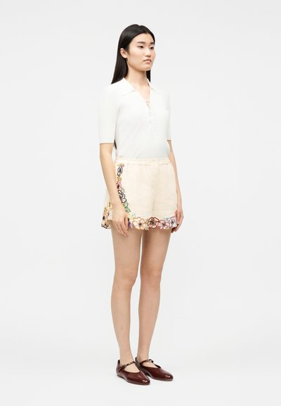 Femme debout portant un haut blanc à manches courtes, un short beige avec une broderie florale colorée, et des chaussures Mary Jane marron foncé.