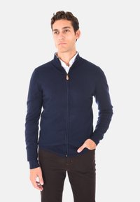 Maglione zip-up navy con collo alto, polsini e orlo a coste. Dotato di un piccolo tirante in pelle per la cerniera. Indossato sopra una camicia bianca con colletto.