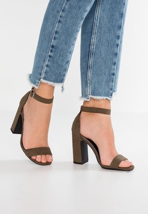 Fødder iført brune ruskind blokhælte sandaler med ankelremme kombineret med lyseblå jeans med frynset kant på en hvid gulv.