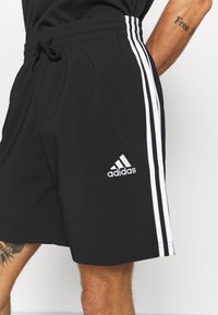 Svarta sportshorts med vitt Adidas-logga och tre vertikala vita ränder. Har en elastisk midja med dragsko.