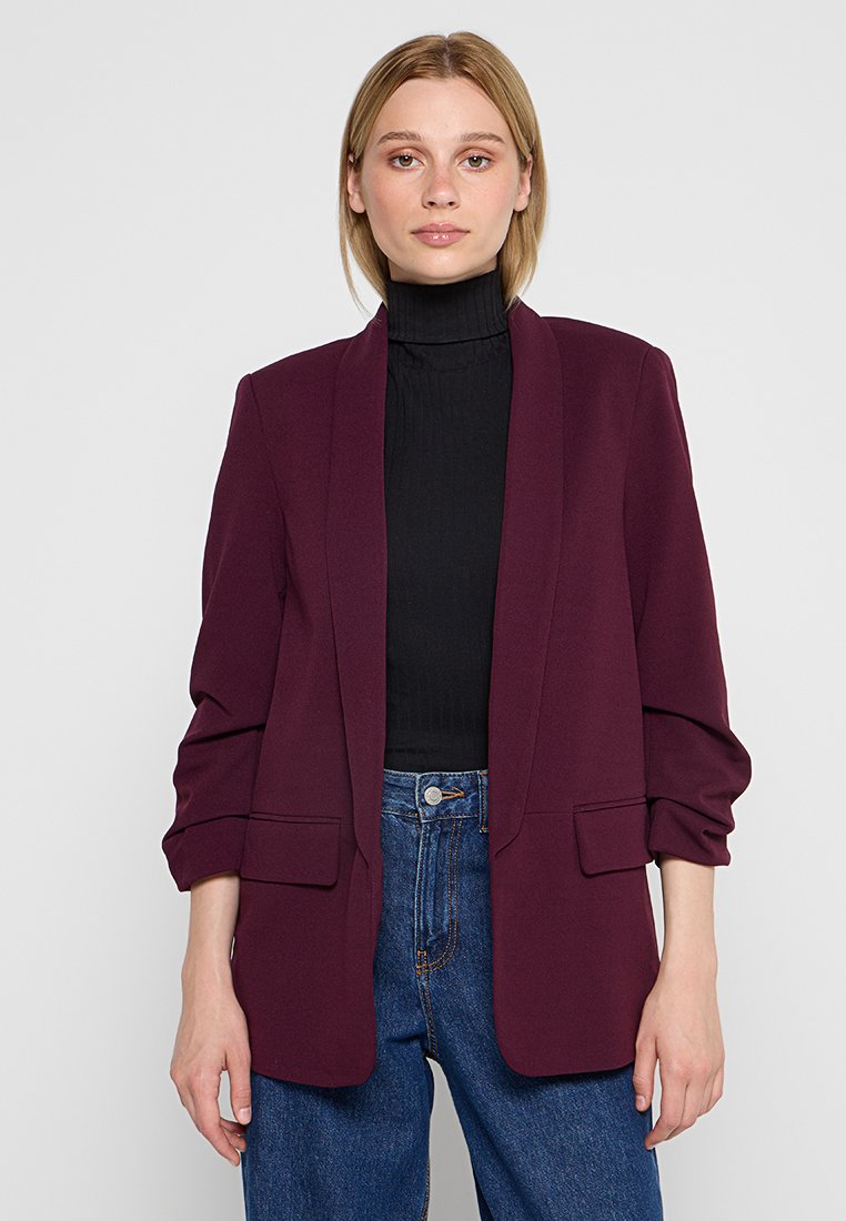 Blazer burdeos con cuello de chal, mangas largas y dos bolsillos frontales, llevado sobre un suéter negro de cuello alto y jeans azules. Textura suave.