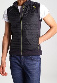 Sort puffet vest med quiltet tekstur, lynlåsefront og grønne accenter. Har sidelommer og en tætsiddende pasform, båret over en hvid T-shirt.