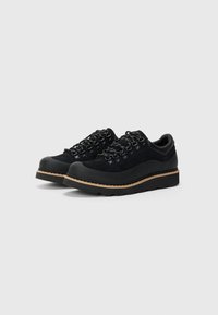 Ein Paar schwarze, knöchelhohe Wanderschuhe mit dicken Sohlen, Metallösen und schwarz-weißen Schnürsenkeln auf einem schlichten hellgrauen Hintergrund.