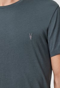 Camiseta gris oscuro hecha de tela suave, con cuello redondo y mangas cortas. Un pequeño diseño de calavera bordado en el pecho.