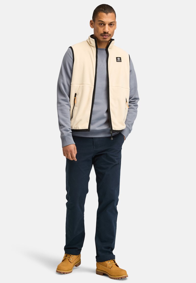 Gilet in pile beige con zip nera, indossato sopra una felpa azzurra chiara, abbinato a pantaloni scuri e stivali gialli. Presenta tasche con zip anteriori.