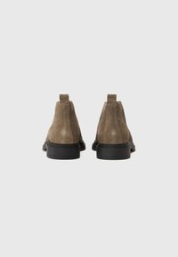 Botines de ante marrón con una textura suave, puntera redondeada y suela plana de goma negra. Presentan un pequeño tirador en la parte posterior.