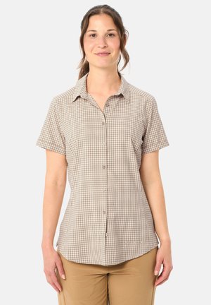 Donna in piedi rivolta in avanti che indossa una camicia a maniche corte beige e bianca a quadri con bottoni e pantaloni marrone chiaro, davanti a uno sfondo semplice.