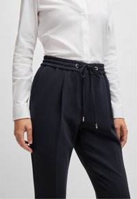 Marinblå byxor i mjukt material, med ett elastiskt midjeband med dragsko och metallaccenter i silverfärg.