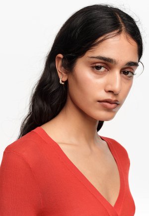Kobieta o długich ciemnych włosach, ubrana w czerwony top z dekoltem w serek i małe złote kolczyki koła, patrząca lekko na bok na białym tle.