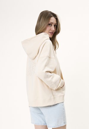 Kvinde med langt hår iført en lysebeige hættetrøje med hætte og lyseblå cowboyshorts, der kigger over skulderen.