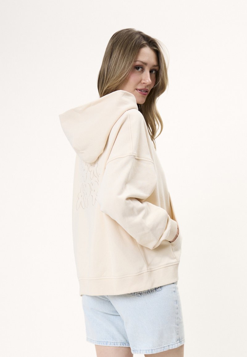 Femme aux longs cheveux portant un sweat à capuche beige clair avec capuche et un short en jean bleu clair, regardant par-dessus son épaule.