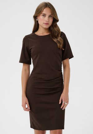 Femme aux cheveux châtain clair portant une robe ajustée marron foncé longueur genoux à manches courtes et des boucles d’oreilles argentées en forme de créoles.