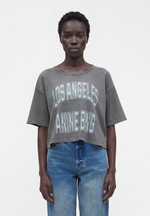 Person stående iført en falmet grå crop top med teksten "LOS ANGELES ANINE BING" og blå jeans mod en ensfarvet hvid baggrund.