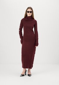 Bordeaux gestricktes Kleid mit hohem Kragen, voluminösen Ärmeln, Knopfdetails entlang eines Arms und einer schlanken Silhouette, die bis zu den Knöcheln reicht.