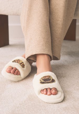 Person trägt beige Hose und weiße flauschige Hausschuhe mit dekorativer Perlenverzierung, steht auf hellem Teppich nahe hölzernen Möbelbeinen.