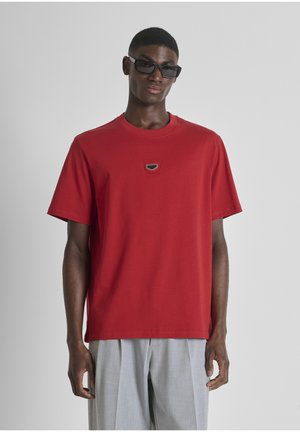 Uomo che indossa occhiali da sole rettangolari neri, una T-shirt rossa a maniche corte con un piccolo logo sul petto e pantaloni grigio chiaro su uno sfondo neutro.