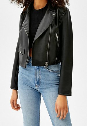 Personne portant une veste en cuir noire courte, un haut en maille noir et un jean bleu clair taille haute, debout devant un fond uni.