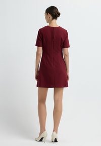 Robe bordeaux à manches courtes avec un col rond, forme ajustée, fermeture éclair au dos et texture lisse ; portée avec des talons hauts de couleur claire.