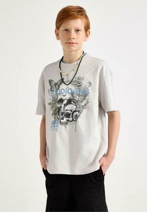 Rødtop dreng iført en lysegrå T-shirt med tryk af kranium, sommerfugle og "STUDIO SK8", sorte shorts og to perlekæder.