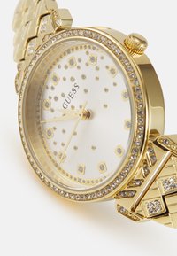 Gouden horloge met een ronde wijzerplaat, crystal accenten en een gepatterde metalen armband. Witte wijzerplaat met gouden details en logo.