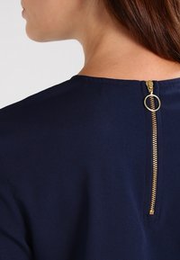 Marinblus med en gulddragkedja på baksidan, rund halsringning och slät textur. Designen är enkel och elegant.