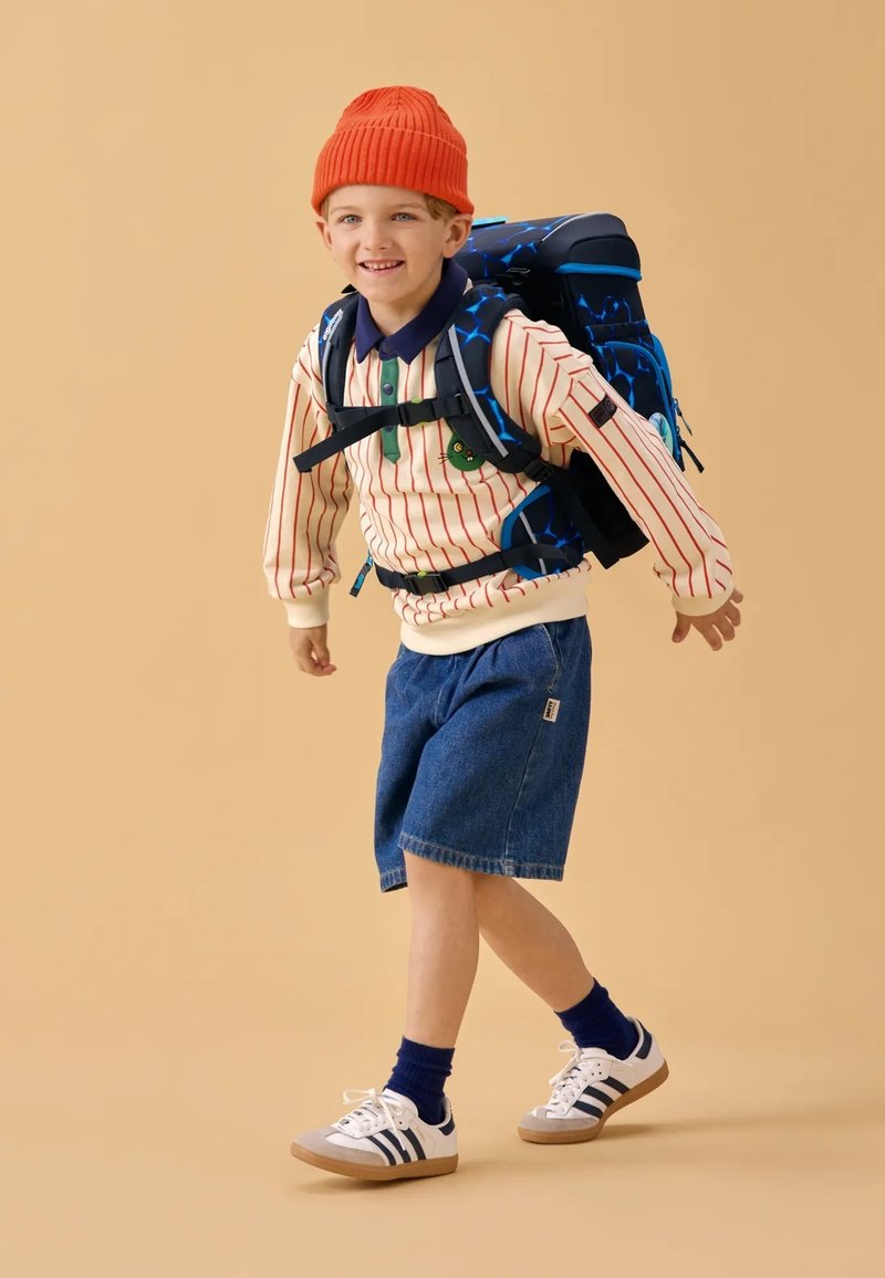 Un sac à dos bleu et noir sur un garçon portant une chemise à rayures à manches longues, un short en denim, un bonnet orange et des baskets blanches avec des accents bleu et noir.