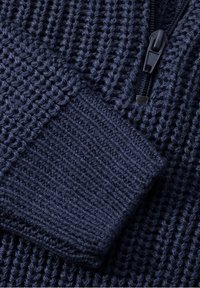 Gros plan d'un tissu tricoté bleu foncé avec un poignet côtelé et une fermeture éclair noire sur un matériau texturé.