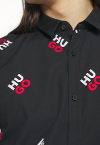 Czarna koszula na guziki z powtarzającym się wzorem słów "HUGO" i "GO" w czerwieni i bieli. Gładka tkanina z usztywnionym kołnierzem.