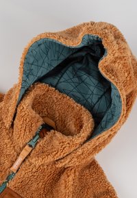 Beige Fleece-Jacke mit einer strukturierten, plüschigen Außenseite und einem blauen gemusterten Innenfutter. Verfügt über eine Kapuze und einen frontalen Reißverschluss.