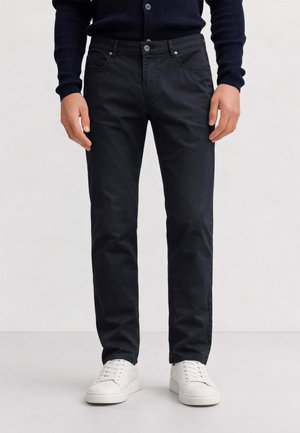 Uomo che indossa jeans neri a taglio dritto, scarpe da ginnastica bianche e un maglione scuro a coste, in piedi davanti a uno sfondo uniforme.