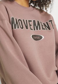 Bordowy, oversize'owy sweatshirt z napisem "MOTION" wydrukowanym dużymi czarnymi literami oraz "1998" w białym owalnym tle. Wykonany z miękkiego materiału bawełnianego, z długimi rękawami.