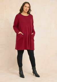 Vrouw staat met handen in de zakken, draagt een burgundy tuniek met lange mouwen, zwarte aansluitende broek en zwarte enkellaarsjes.