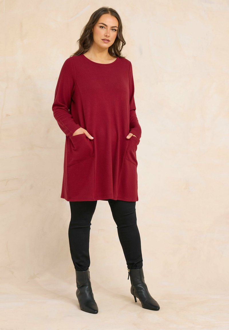 Vrouw staat met handen in de zakken, draagt een burgundy tuniek met lange mouwen, zwarte aansluitende broek en zwarte enkellaarsjes.
