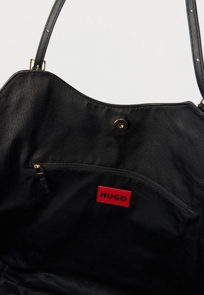 Intérieur de sac à main noir avec une poche zippée, une étiquette rouge HUGO, et une fermeture magnétique sous la fine bandoulière noire.