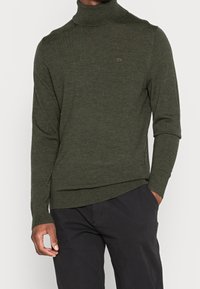 Uomo che indossa un maglione a collo alto verde oliva con il logo CK sul petto e pantaloni neri, con una mano in tasca su uno sfondo bianco.