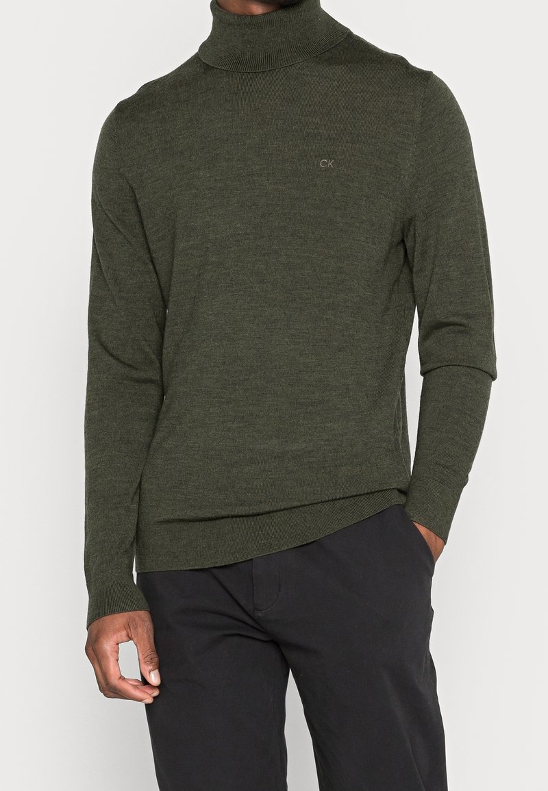 Uomo che indossa un maglione a collo alto verde oliva con il logo CK sul petto e pantaloni neri, con una mano in tasca su uno sfondo bianco.