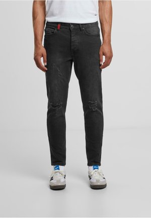 Black denim jeans with a slim fit, distressed details, five pockets, and a red accent on the waistband, paired with white sneakers.