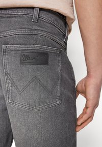 Harmaat denim-farkut, joissa on nahkainen Wrangler-tagi, erottuva chevron-ommeltu takatasku ja metallirivettejä saumoissa.
