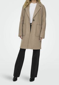 Abrigo de mezcla de lana beige con un solo botón, solapas con muesca y dos bolsillos frontales de parche, combinado con jeans negros de pierna ancha y botas de tacón.