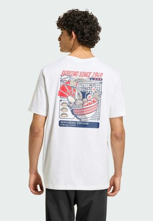 Mand med krøllet hår iført en hvid T-shirt med et farverigt billede af en ramen-skål og rød tekst "Serving Since 1949" på bagsiden.