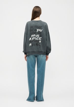 Persona con capelli castani lunghi che indossa una felpa oversize grigio scuro con testo bianco e nero e jeans azzurri, in piedi di spalle.