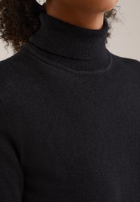 Une personne portant un pull à col roulé noir, montrant le cou et la partie supérieure de la poitrine avec des cheveux frisés partiellement visibles.