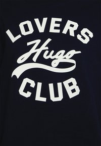 Camiseta azul marino con texto blanco audaz y contrastante que dice "LOVERS HUGO CLUB" en una mezcla de fuentes en mayúsculas y cursiva.