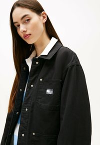 Chaqueta de utilidad negra con botones a presión, un bolsillo en el pecho con un parche de logo, y una camisa blanca de cuello debajo. Textura de tela suave.