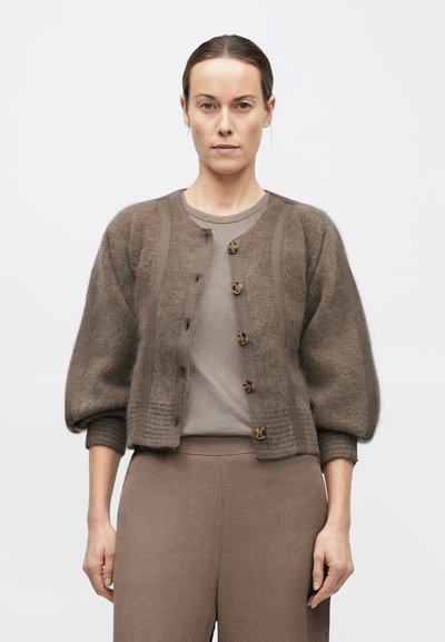 Femeie purtând un pulover taupe cu dungi, un cardigan scurt din material pufos cu nasturi în model tartaruga și pantaloni largi cu talie înaltă, stând pe un fundal simplu.