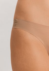 String beige en tissu extensible doux. Texture lisse avec une fine ceinture élastique, conçu pour une couverture minimale et un confort optimal.