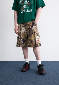 Πράσινη δικτυωτή μπλούζα Adidas με λογότυπα; καφέ καμουφλάζ cargo σορτς; μαύρα και κόκκινα αθλητικά παπούτσια; λευκές κοντές κάλτσες; όρθιος σε μπλε χαλί.